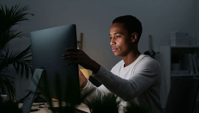 Focused young professional adjusting large monitor in dim home office late-night work with warm glow
