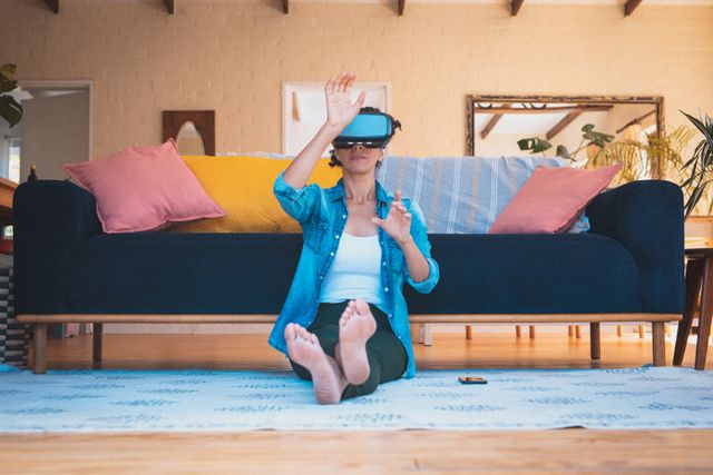 Woman sitting on floor in living room using VR headset. Ideal for illustrating modern technology use, home entertainment, and quarantine activities. Suitable for articles on virtual reality, tech trends, and home lifestyle.