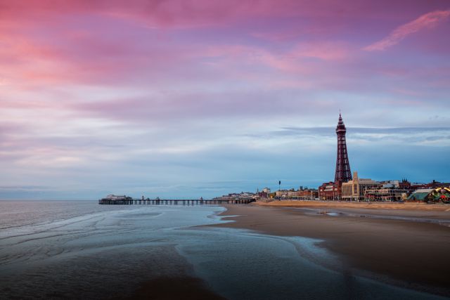Showing Blackpool beach at dawn with a calm, serene atmosphere. The iconic Blackpool Tower and pier are in the distance, bathed in early morning light. The sky features pink and blue hues which add to the tranquility of the scene. This tranquil coastal scene is perfect for travel blogs, tourism promotions, and posters highlighting the beauty of Blackpool and the United Kingdom.