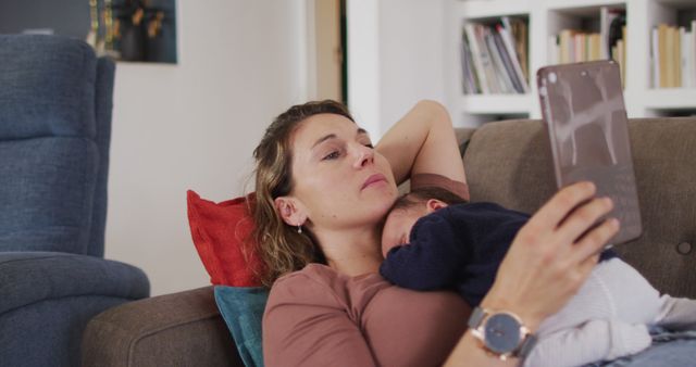 Mother is seen lying on the couch, holding her baby while using a tablet. Ideal for illustrating themes of motherhood, multitasking, modern technology, and home life. Useful for content related to family dynamics, parenting tips, work-from-home scenarios, and technology in daily routines.