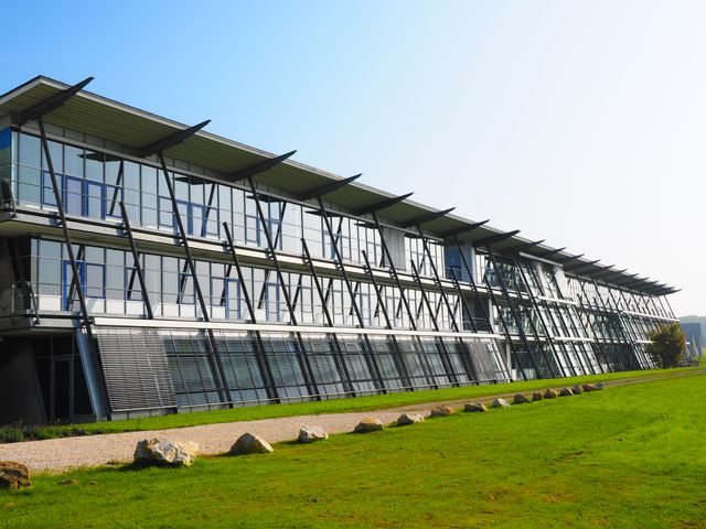 Modern office building featuring large glass windows and a steel structure, situated in a landscaped area with green grass. Sunlight illuminates the facade, creating a clean and professional look. Ideal for advertisements, business brochures, real estate listings or website headers promoting modern architecture or corporate environments.