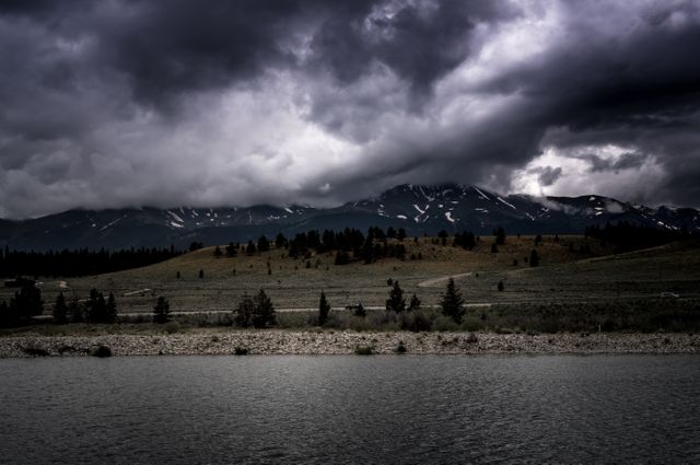 Showcases a dramatic dark sky with heavy clouds over a mountainous area and calm water, ideal for depicting stormy weather, natural beauty, or wilderness adventures. Can be used for travel magazines, weather reports, nature blogs, desktop wallpapers, and relaxation visuals.