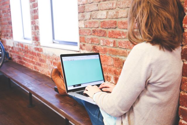This image shows a woman working on a laptop in a loft-style office with exposed brick walls. She sits casually on a wooden bench, engaging with her tasks. This photo can be used in designs and advertisements related to modern workspaces, freelance work, technology use, and productive environments.