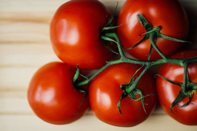 Close-up view of fresh, ripe red tomatoes attached to a vine laying on a light wooden surface. Ideal for use in food blogs, health articles, cooking recipes, restaurant menus, or agricultural content promoting local produce and organic farming.