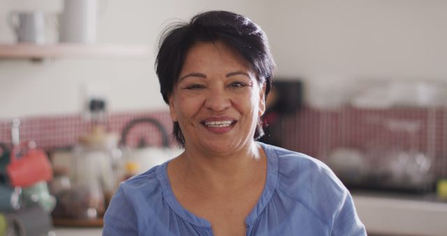 Captures a middle-aged woman smiling warmly in a kitchen, wearing a blue blouse. Perfect for use in lifestyle blogs, articles about domestic life, family-oriented content, or advertisements featuring relatable, everyday scenarios. The warm and candid feel adds authenticity and emotional connection.