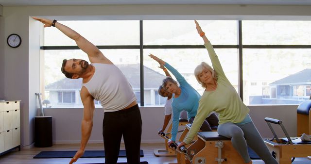 A diverse group consisting of a middle-aged Caucasian man and two senior women are engaged in a stretching exercise at a Pilates studio, with copy space. Their coordinated movement suggests a focus on flexibility and wellness in a supportive fitness environment.