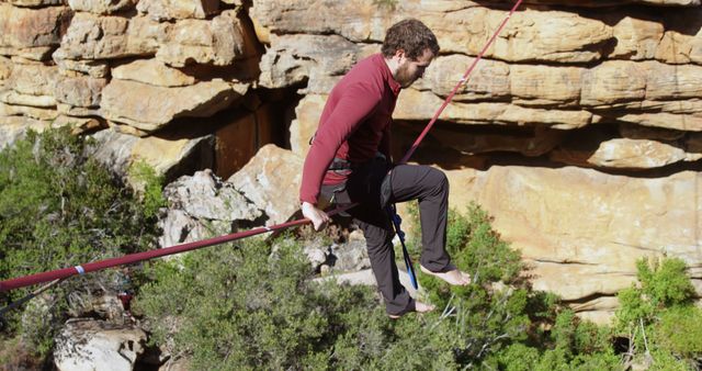 Man balancing with focus on slackline in rocky, outdoor environment. Suitable for themes of outdoor adventure, extreme sports, and nature exploration. Ideal for illustrating physical agility, concentration, and the challenge of outdoor activities in rugged terrain.