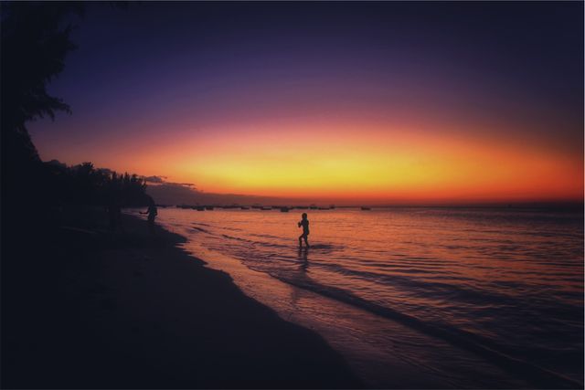 Image depicts silhouette of person walking along tranquil beach during sunset. Ideal for travel blogs, relaxation and serenity themes, inspirational posters, and nature-related content. Perfect for evoking feelings of calm and peacefulness, highlighting the beauty of twilight at the beach.