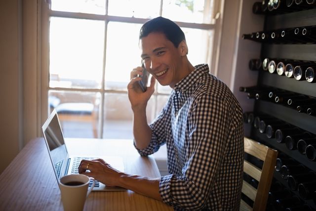 Young man smiling while using laptop and talking on mobile phone in a restaurant. Ideal for depicting modern business communication, remote work, and casual professional settings. Suitable for articles on multitasking, technology in daily life, and work-life balance.
