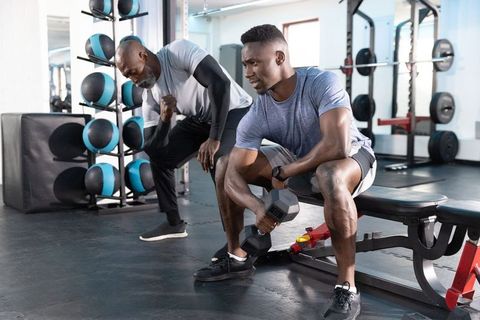 African american trainer coaching male client doing seated dumbbell curl in modern gym