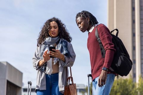 Multiethnic friends checking smartphone with luggage on urban campus plaza