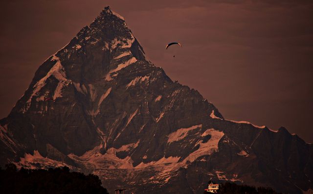 Paraglider flying near towering, majestic mountain with snow-capped peaks during dusk. Perfect for promoting adventure travel, excitement of extreme sports, and breathtaking natural landscapes. Ideal for magazines, travel brochures, and websites focusing on outdoor tourism.