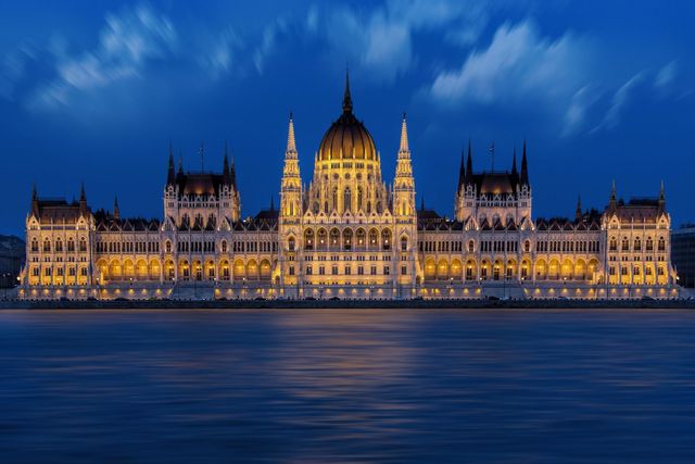 Hungarian Parliament Building glowing against night sky, reflecting in calm waters of Danube River. Gothic Revival architecture with illuminated domes and spires. Perfect for travel guides, historical features, tourism brochures, and architectural studies focusing on Budapest's landmarks.