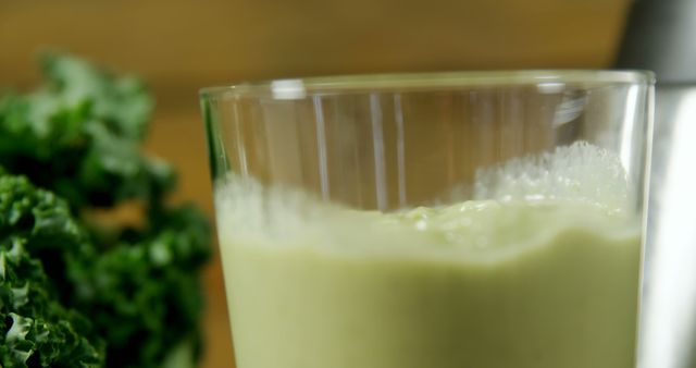 A close-up view of a glass of green smoothie, with fresh kale visible in the background, with copy space. Green smoothies are popular for their health benefits and are often included in fitness and wellness routines.