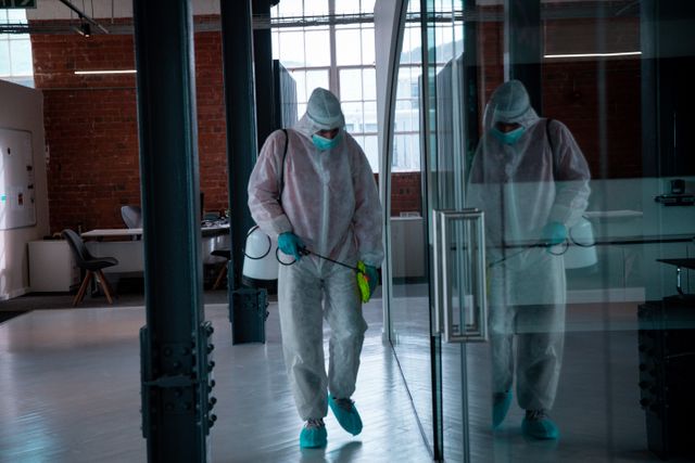 Worker in protective coverall disinfecting modern office space during COVID-19 pandemic. Useful for illustrating workplace safety, hygiene practices, and pandemic-related precautions in office environments.