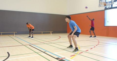 Diverse men serving and defending volleyball on indoor hardwood court, teammates ready