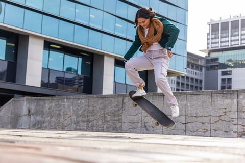 Young woman performing kickflip on skateboard in modern city plaza with glass office buildings