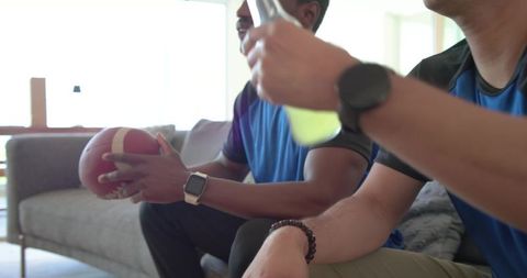 Diverse male friends sitting on sofa watching game while holding football and drink