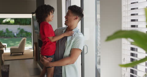 Father Carrying Son Smiles Joyfully at Home by Window
