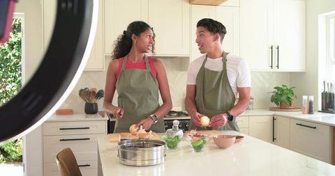 Young couple wearing olive aprons filming cooking tutorial at kitchen island with ring light