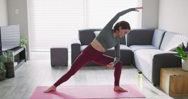 Young woman practicing yoga poses on pink mat in modern living room. Ideal for promoting home exercise routines, healthy lifestyles, and indoor fitness practice. Great for use in fitness blogs, yoga class promotions, home workout tutorials, and social media posts reflecting wellness and relaxation.