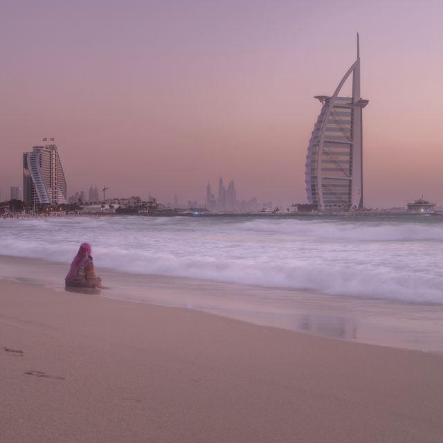 Person sits alone on Dubai beach during sunset, with Burj Al Arab in background. Perfect for topics on travel, meditation, solitude, hospitality industry, and Middle Eastern tourism.