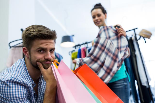 Man holding colorful shopping bags while woman looks at plaid shirt in clothing store. Ideal for illustrating shopping experiences, retail therapy, consumer behavior, and relationship dynamics during shopping trips.