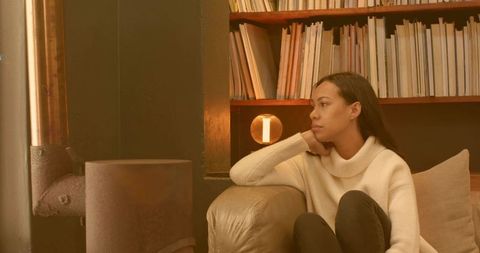 Pensive woman relaxing on cozy sofa beside bookshelf and wood stove in warm amber living room