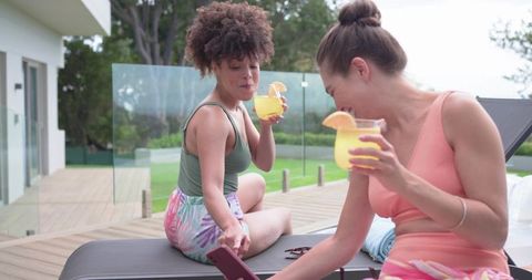Two friends relaxing on poolside deck sipping citrus cocktails and sharing phone laughs
