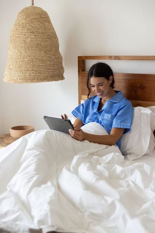 Woman in blue pajamas relaxing in bed with tablet and white duvet