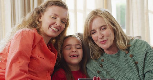 Three women from different generations sitting together on sofa smiling while looking at a tablet. Useful for promotions on family bonding, technology use across ages, home life, and multigenerational living.