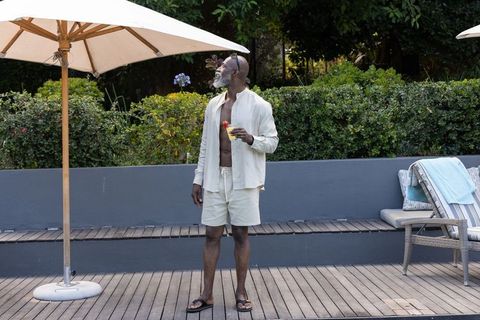 African american man enjoying cocktail on pool deck under umbrella, summer lifestyle