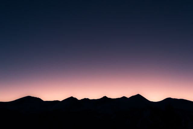 Silhouettes of mountains stand against a vibrant gradient sky transitioning from deep blue to soft pink. Peaceful and serene image perfect for use in travel blogs, landscape photography, backgrounds, desktop wallpapers, posters, nature-themed advertisements, and more. Highlights natural landscapes during twilight hours, depicting the serene beauty of dawn or dusk in a mountainous region.