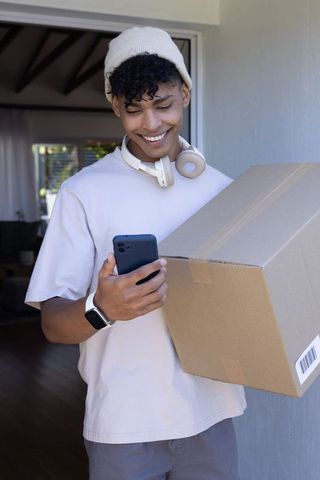 Young african american man holding parcel while checking smartphone at front door smiling