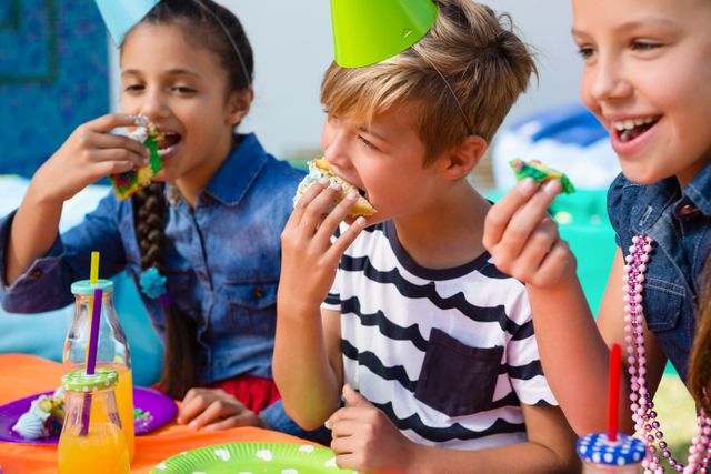 Children are sitting at a table, enjoying cake and drinks during a birthday party. They are wearing party hats and smiling, creating a joyful and festive atmosphere. This image can be used for advertisements, party planning websites, or any content related to children's celebrations and events.