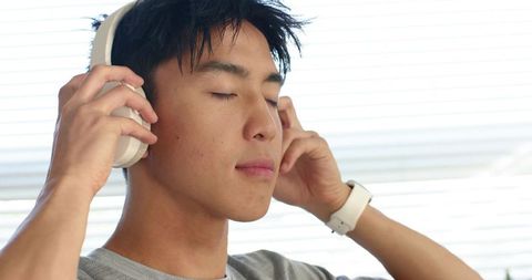 Asian man listening with white headphones, adjusting headset, calm focused modern lifestyle