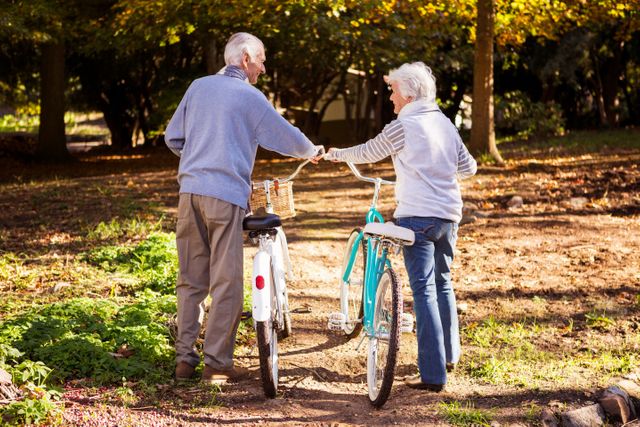 Senior couple walking side by side with their bicycles in a park during autumn. Ideal for use in advertisements promoting healthy lifestyles, retirement living, outdoor activities, and senior companionship. Can be used in brochures, websites, and social media posts focused on active aging and wellness.