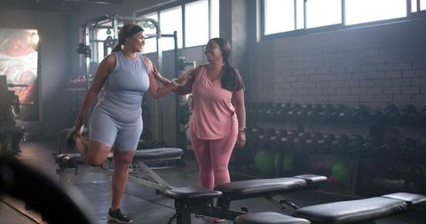 Diverse women stretching and encouraging each other during gym workout near dumbbell rack