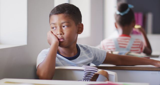 Image of thoughtful biracial boy sitting at desk in classroom. primary school education and learning concept.