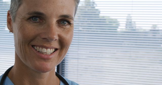 This image shows a close-up of a mature female doctor smiling inside her medical office. Perfect for use in healthcare promotions, advertisements for medical services, websites for doctor profiles, or materials promoting healthcare. The doctor's friendly expression emphasizes trust and professionalism.