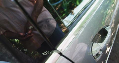 Man leaning on silver car door holding smartphone with reflected numeric codes on glass