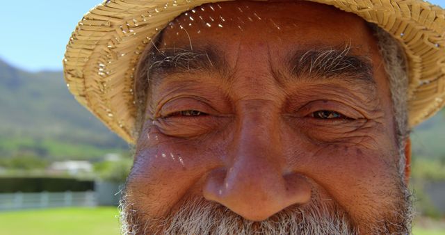 Elderly man smiling outdoors, wearing a straw sun hat, enjoying sunny day. Suitable for depicting happiness, aging gracefully, outdoor activities, summer lifestyle, and relaxation.