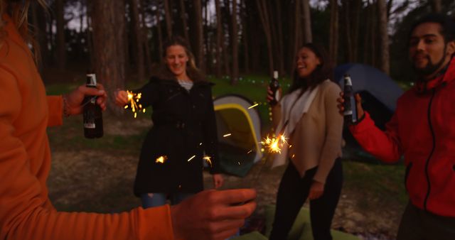 A group of friends is enjoying a night outdoors in a forest. They are celebrating and having fun with sparklers and drinks in hand while surrounded by tents. This scene can be used for promoting outdoor activities, camping gear, or summer festivities and friendship.