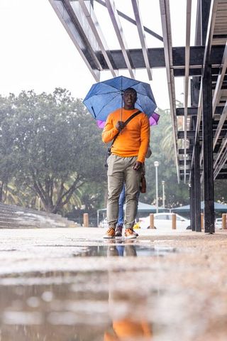 Walking through rainy plaza holding blue umbrella and orange sweater reflecting in puddle
