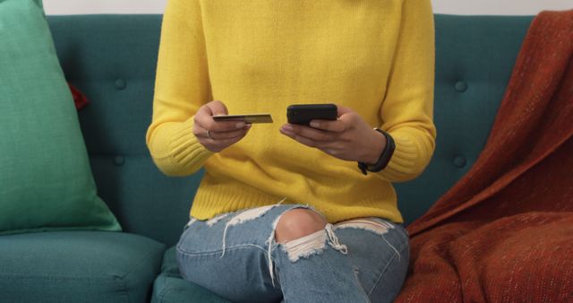 Woman wearing yellow sweater and jeans using credit card while holding mobile phone, symbolizing online shopping and digital payment. Ideal for illustrating e-commerce convenience, secure transactions, modern technology, and shopping apps. Perfect for advertisements, blog articles on online shopping benefits, and promotional content for e-commerce platforms.