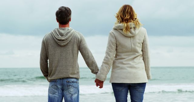 A Caucasian man and woman are holding hands while looking out at the ocean, with copy space. Their relaxed posture and casual clothing suggest a moment of peaceful connection with nature.