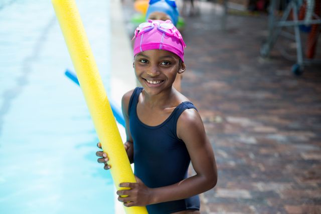 Young girl in swimwear holding a yellow pool noodle while standing at the edge of a swimming pool. She is wearing a pink swim cap and smiling. Ideal for use in advertisements for swimming lessons, summer camps, children's activities, and recreational programs.