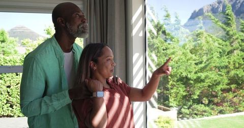 Diverse couple admiring mountain view through sliding glass door, pointing at garden