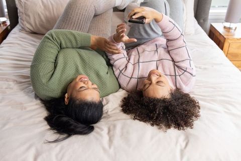 Diverse young women laughing and scrolling smartphone while lying on bed in cozy sweaters