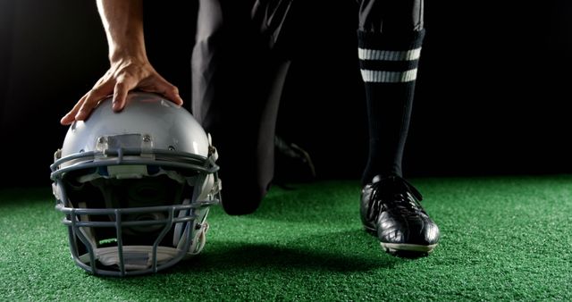 Football player kneeling on turf field, touching helmet with hand. Ideal for sports-related content, athletic gear ads, or motivational material.